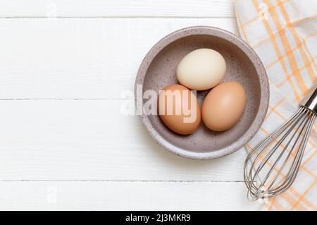 Three chicken eggs in a bowl on a white wooden background. Top view. Ingredients for breakfast, baking, mayonnaise. Stock Photo