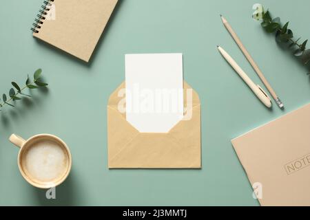 Envelope with blank card inside, cup of coffee, paper notepad, weekly planner, eucalyptus leaves on green table. Flat lay, top view, copy space. Stock Photo