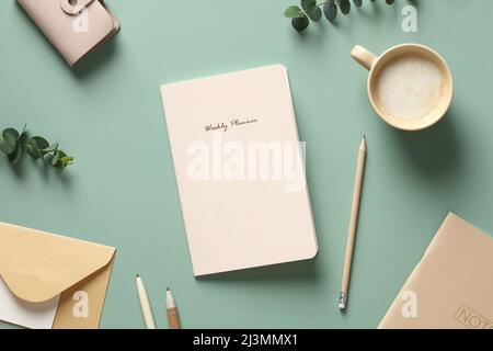 Flat lay weekly planner, cup of coffee, pencil, envelope, paper notebook, accessories on green table. Home office desk, feminine workspace. Stock Photo