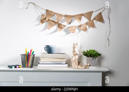 Echeveria in geometric pot, stack of books, pens in a pencil holder, felt-tip pens, wooden figurine of a hare are on shelf. Garland of craft paper fla Stock Photo