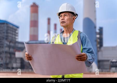 Senior supervisor engineer in his work cloths holding construction blueprints to check on the project progress at the power plant site, blurred power Stock Photo