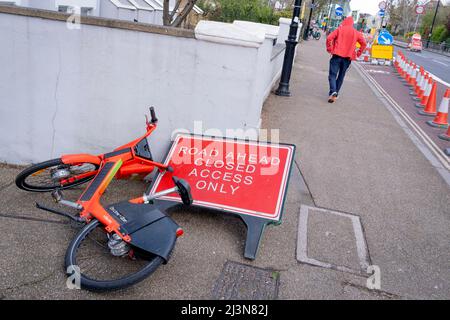 During a windy day in the capital, a red Santander rental bike has fallen over a road sign near Hammersmith Bridge, now closed to motor traffic but open to pedestrians, 6th April 2022, in London, England. Stock Photo