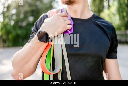 Sportive man guy holding in his hand wearing fitness tracker set of colorful elastic rubber bands at the stadium outdoors. Athlete male person during