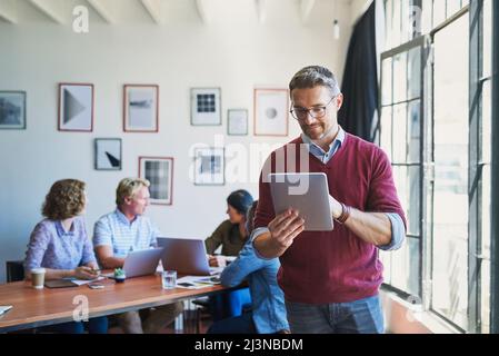 The digital manager gets things done. Shot of a mature man using a digital tablet with his team in the background of a modern office. Stock Photo