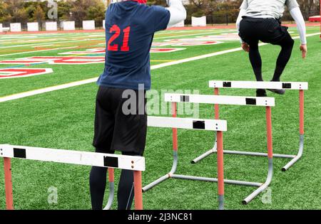 Rear view of two high school boys jumping over track hurdles on a green turf field during strength and agility practice. Stock Photo