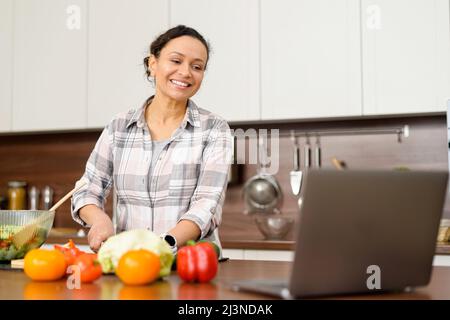 Cooking at home. Mid-age female is watching cooking video recipes on a laptop and cutting salad. Housewife preparing food and watching TV series Stock Photo