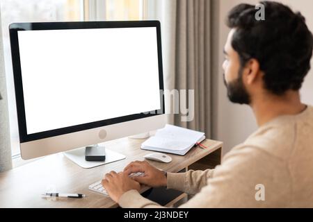 Back view of positive Hindi man in smart casual clothes using computer while sitting at the desk in his flat. Young Indian male student watching webinars, educational courses, learning on the distance Stock Photo