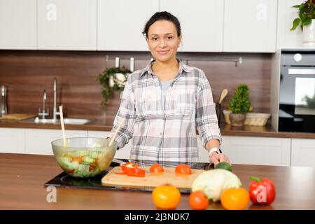 Smiling middle-aged woman in casual shirt preparing lunch in the kitchen. Housewife going to make salad, stands near countertop and looks at camera, vegetable on the table. Healthy lifestyle concept Stock Photo