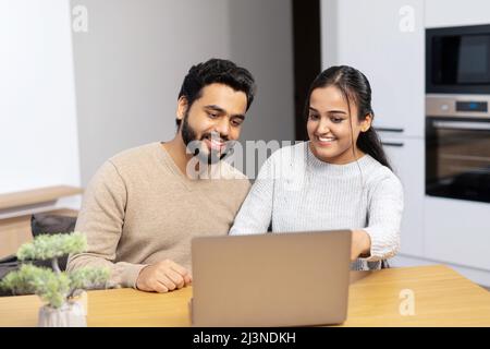 Happy woman and man in embrace look at the screen and laughs, watching videos, video calling with friend. Cute middle eastern couple enjoys leisure time at home with a laptop Stock Photo