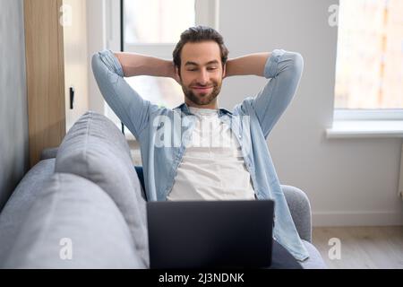 Handsome young European man with beard sitting on the sofa with laptop at home, searching in the internet, watching film or serial, remote meeting, on the weekend, relaxed, listening performance Stock Photo
