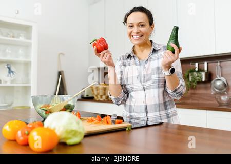 Smiling middle-aged woman going to cooking vegetable meals, standing in the modern kitchen, holding avocado and pepper in hands, looking at camera Stock Photo