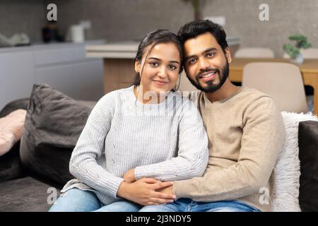 Multi ethnic couple in love rent a house together. Happy Indian newlyweds moved in new apartment, sitting at the sofa in embraces, looking at the camera Stock Photo