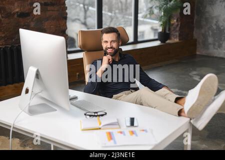 Smiling caucasian man wearing casual wear sitting at the armchair with his legs at the table and using computer at modern office. Confident male entrepreneur concept Stock Photo