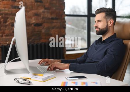 Focused and smiled man in smart casual shirt looking at laptop screen. Cheerful caucasian guy sitting at the desk and solving his daily tasks Stock Photo