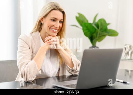 Smiling young caucasian female entrepreneur sitting at the table with laptop, working from home and looking at the screen, watching an online webinar or a course, typing email Stock Photo