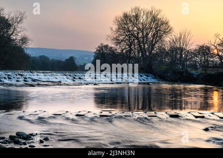 Scenic rural landscape (water flowing over weir steps, stepping stones crossing place) - River Wharfe, Burley in Wharfedale, Yorkshire, England, UK. Stock Photo