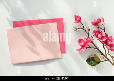 Blank postcard with envelope. Pink decorative magnolia flowers. Top view on off white background. Natural direct sunlight, shadows. Stock Photo