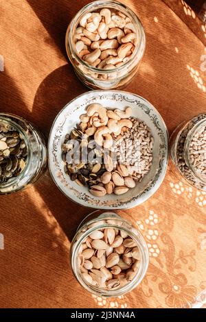 Cashew nuts, pistachios, sunflower seeds and pumpkin seeds in a glass jars and porcelain bowl on the table. Healthy snacks Stock Photo