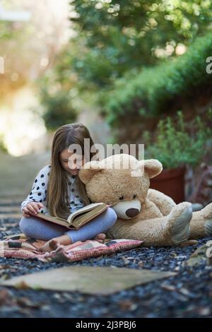 Teddy seems to be taking a nap from storytime. Shot of a little girl reading a book with her teddy bear beside her. Stock Photo