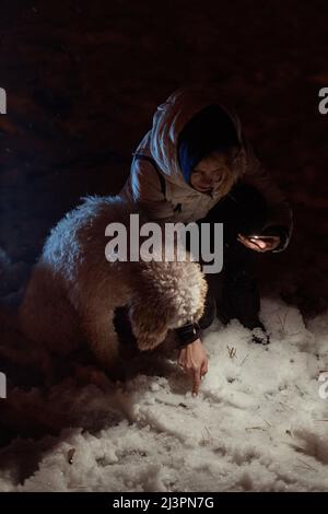 A woman with a dog is looking for a lost thing by the light of a flashlight outside in the winter. Stock Photo