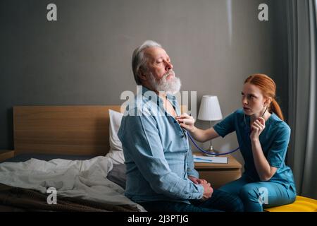 Female doctor listening with stethoscope lungs and heart of senior aged male patient sitting on bed at home. Stock Photo
