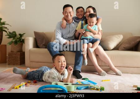 Little boy lying on floor and sticking out tongue when his parents and siblings sitting on sofa and hugging Stock Photo