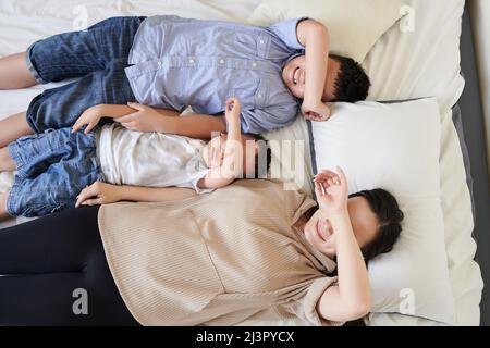 Smiling mother and two sons lying on bed and covering eyes with arms, view from above Stock Photo