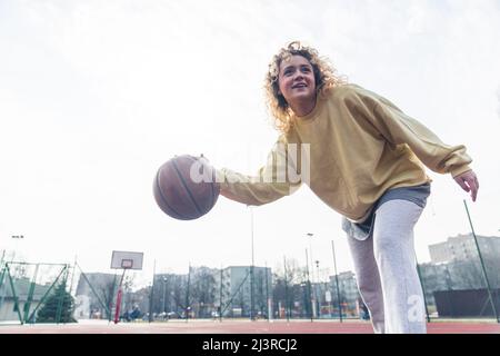 Excited young blond woman plays basketball in baggy clothes on the sports field, looking up copy space medium full shot . High quality photo Stock Photo