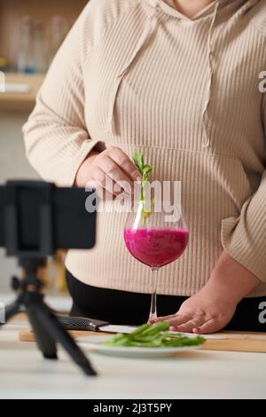 Plus-size female influencer filming herself putting arugula greens in glass of red smoothie Stock Photo