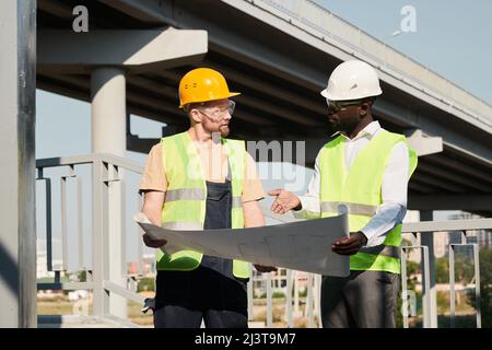 African-American architect in white hardhat and vest pointing at blueprint while working with builder at construction site Stock Photo