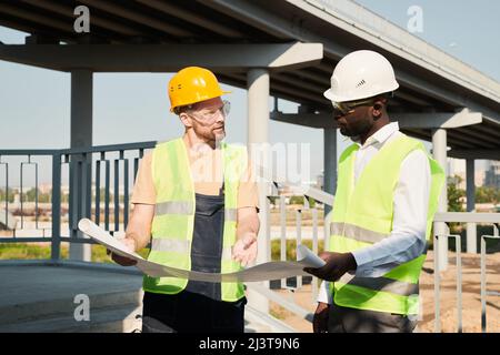 Young African-American construction manager in hardhat and glasses holding blueprint and listening to foreman at building site Stock Photo