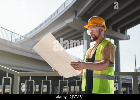 Construction worker examining blueprint and standing on building site of apartment house, large block of flats under construction Stock Photo