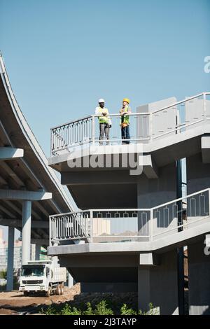 Engineers in white hardhats and safety work vests discussing building project while viewing blueprint at construction site of apartment building Stock Photo