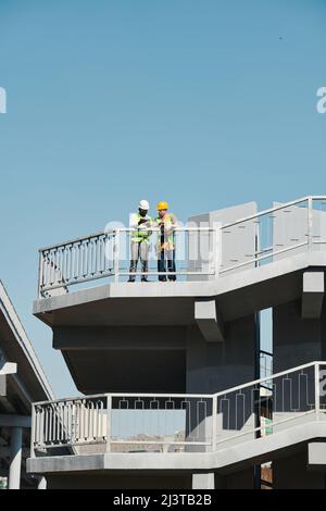 Confident young mixed race construction engineer in eyeglasses talking to coworker and gesturing while explaining plan of work to him Stock Photo