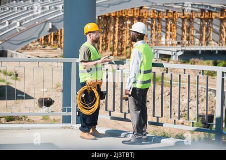 Engineers in hardhats and green vests discussing construction methods while viewing blueprint and standing on construction site with frames and cables Stock Photo