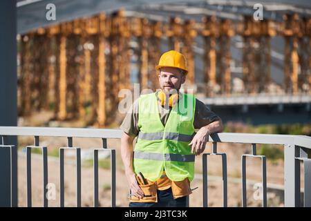 Waist up portrait of cheerful Middle-Eastern construction worker wearing harhat and holding metal bars smiling at camera while enjoying work on site Stock Photo