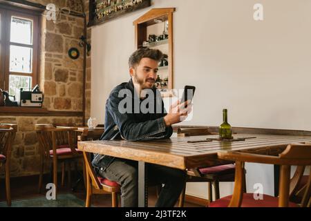 Young man puts his arms on a wooden table and looks at the phone in his hand, an old cafe and a nostalgic look, wood tones and aesthetic place Stock Photo