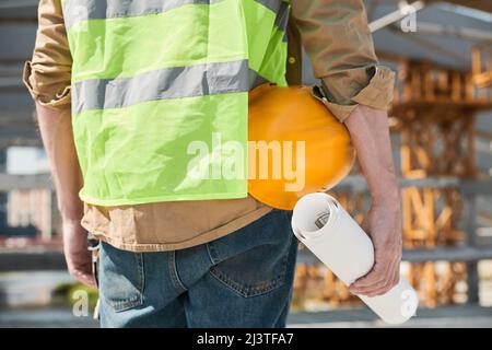 Unknown construction workers holding blueprint and hardhats walking across site outdoors, copy space Stock Photo