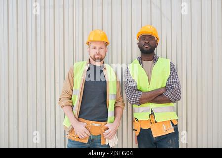 Builders in safety vests and hardhats sitting on cement beam and talking while relaxing on construction site during coffee break Stock Photo