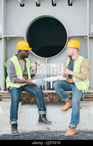 Content young multi-ethnic construction coworkers sitting on concrete beam with steel bars and chatting while drinking coffee during break Stock Photo