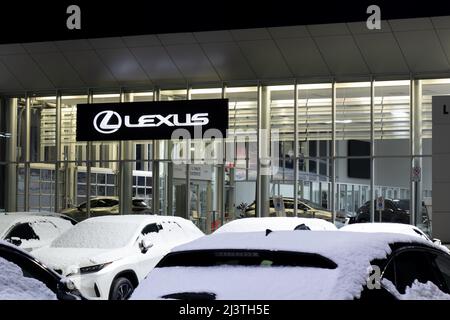 The Lexus logo on a hanging sign at the front of a Lexus dealership at night, seen from behind a parked new car. Stock Photo