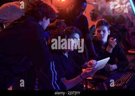 Young curly-haired man in headphones using tablet while explaining strategy of online game to friends in computer club Stock Photo