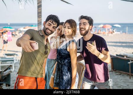 Young people get together taking selfie on the beach. Friends using smartphone and have fun together with the sea in the background Stock Photo