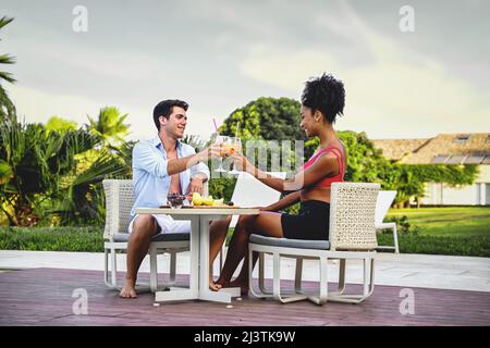 Joyful couple of vacation having breakfast sitting on a table in the terrace - newlyweds toast fruit cocktails at the luxury resort patio Stock Photo