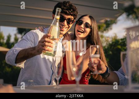 Couple of sweethearts standing on the table pouring sparkling wine champagne into their friends flute glasses celebrating the engaging party outdoors Stock Photo
