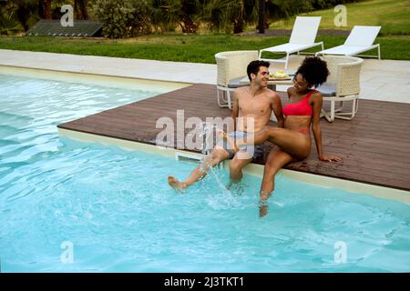 Interracial couple play splash water sitting poolside - multiracial couple sitting by the luxury resort swimming pool Stock Photo