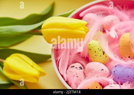 Festive Easter background. Pink bowl with colorful eggs, pink feathers and yellow tulips. Happy Easter concept. Post card on yellow background Stock Photo