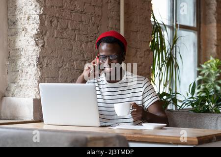 Smiling African man talking on phone, holding cup of coffee, remotely online work on laptop in cafe Stock Photo