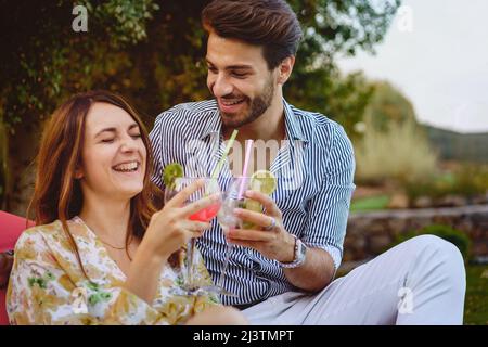 Couple of young people having fun toasting cocktails glasses sitting in the luxury resort garden outdoors Stock Photo