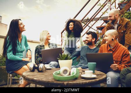 Group of men and women collaborate talking together in a common work space sitting around a table in the university terrace. Stock Photo